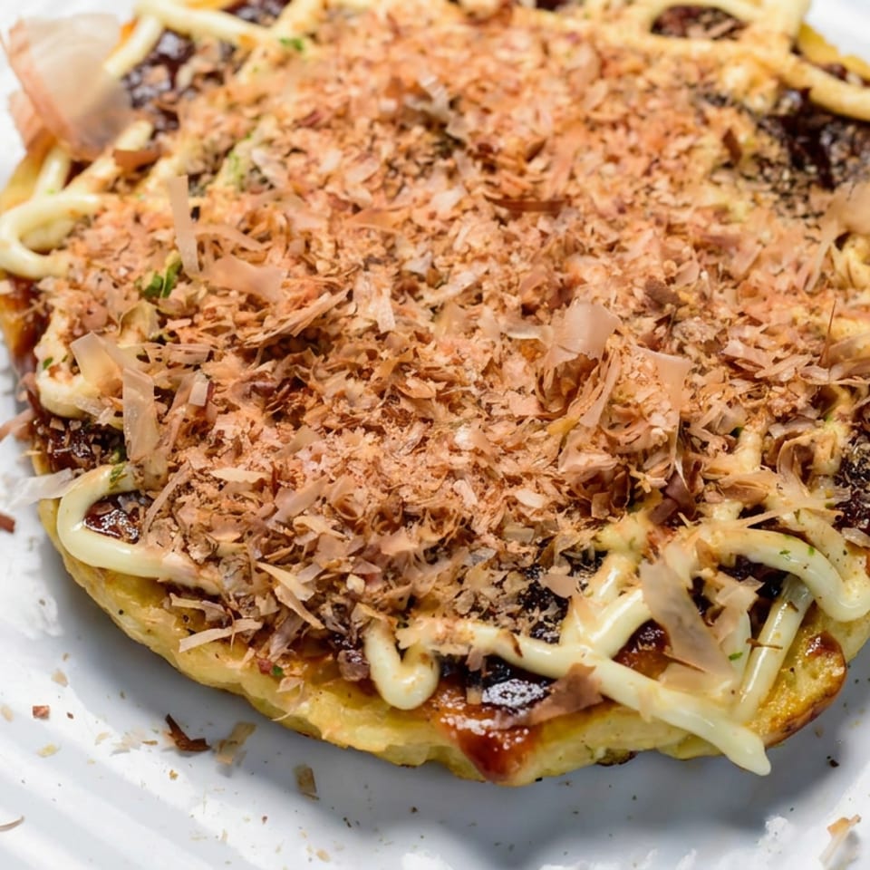 Close-up of Japanese okonomiyaki topped with mayonnaise, savory sauce, and delicate bonito flakes on a white plate.