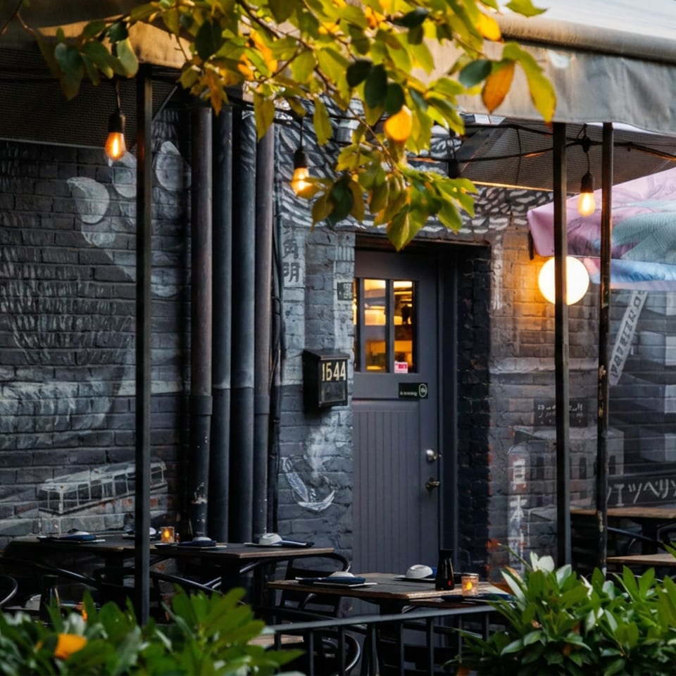 Outdoor restaurant patio with set tables, hanging lights, lush greenery, and a dark brick wall featuring a large mural.