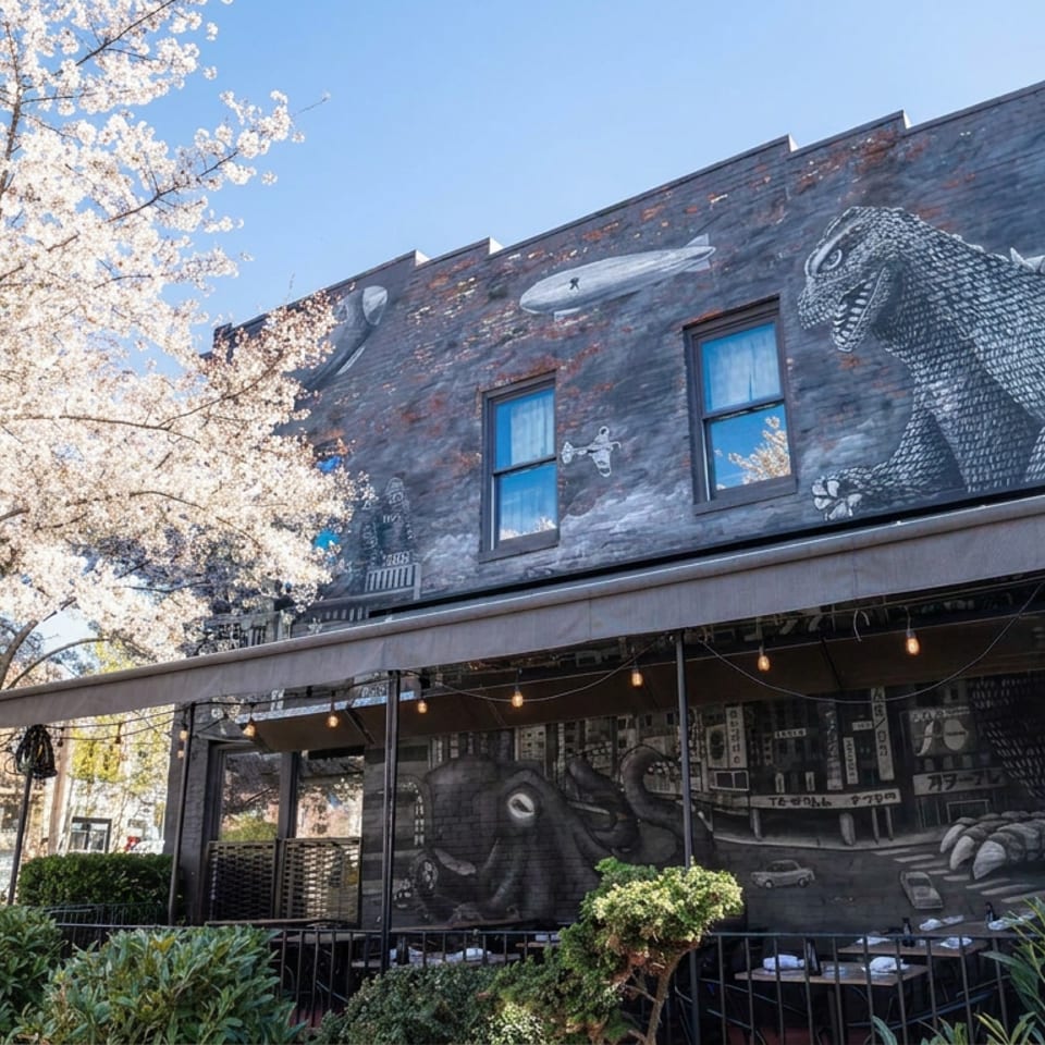 Restaurant storefront with dinosaur murals, outdoor seating, string lights, and a blooming tree under a clear blue sky.