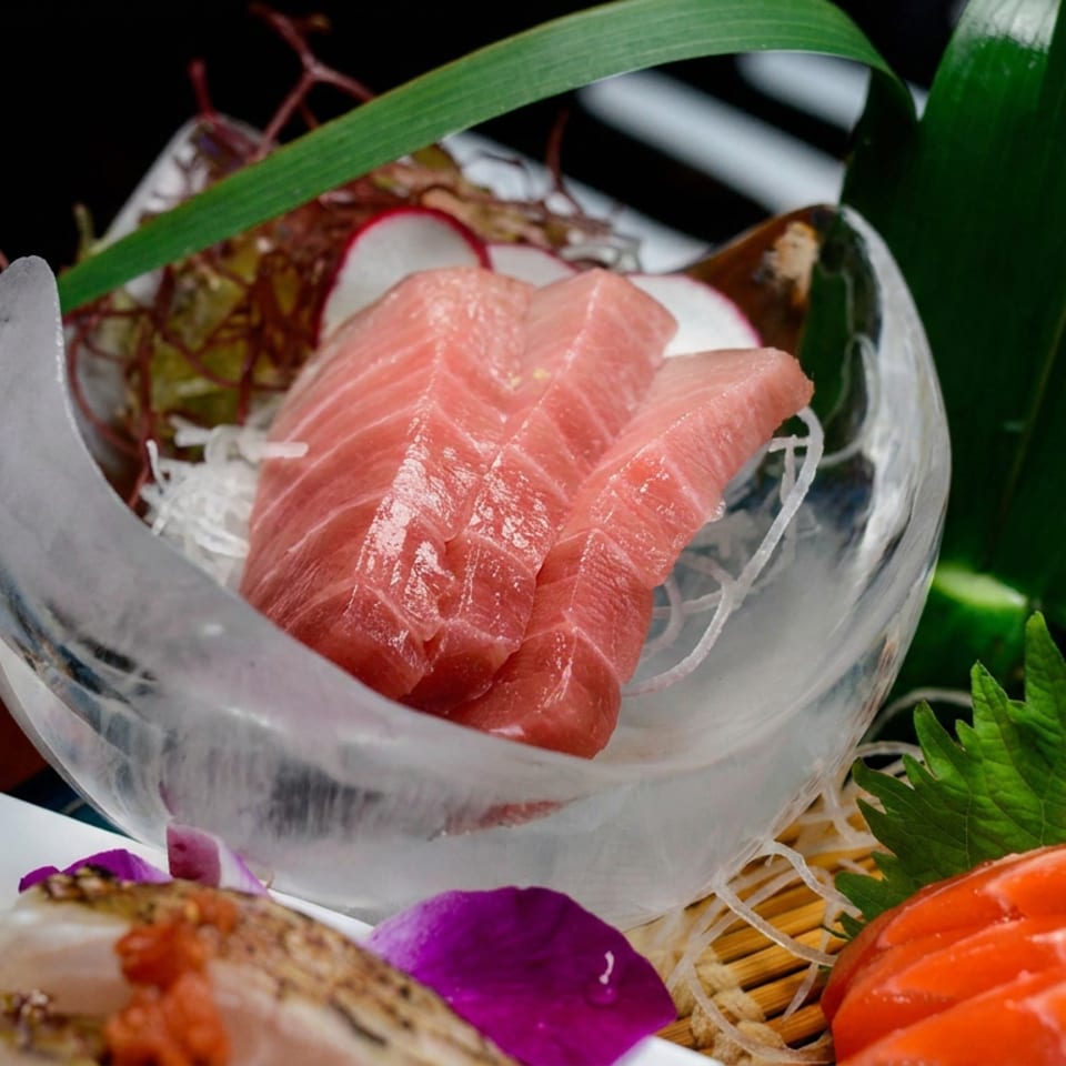 Assorted sashimi featuring pink tuna in a glass bowl, salmon slices, shredded daikon, shiso leaves, and decorative garnishes.