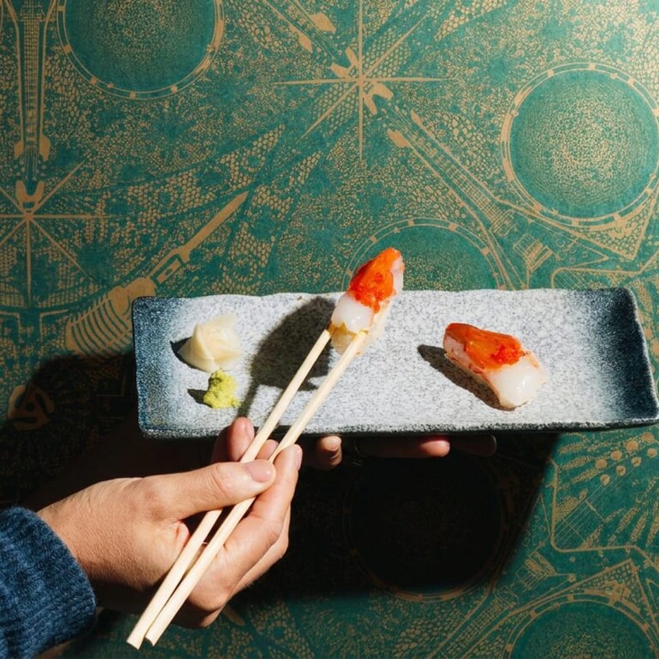 Hand uses chopsticks to lift sushi from a rectangular plate beside wasabi and pickled ginger on a patterned tabletop.
