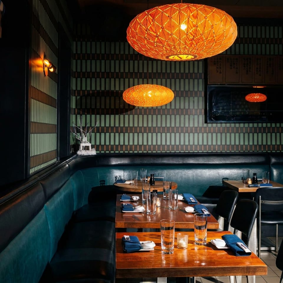 Dimly lit restaurant dining room with teal leather booths, wooden tables, patterned walls, and glowing woven pendant lights.