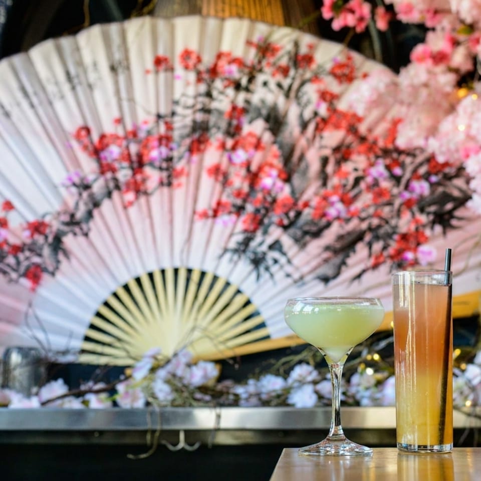 Two colorful cocktails sit on a wooden table before a large floral Japanese fan and pink cherry blossom decorations.