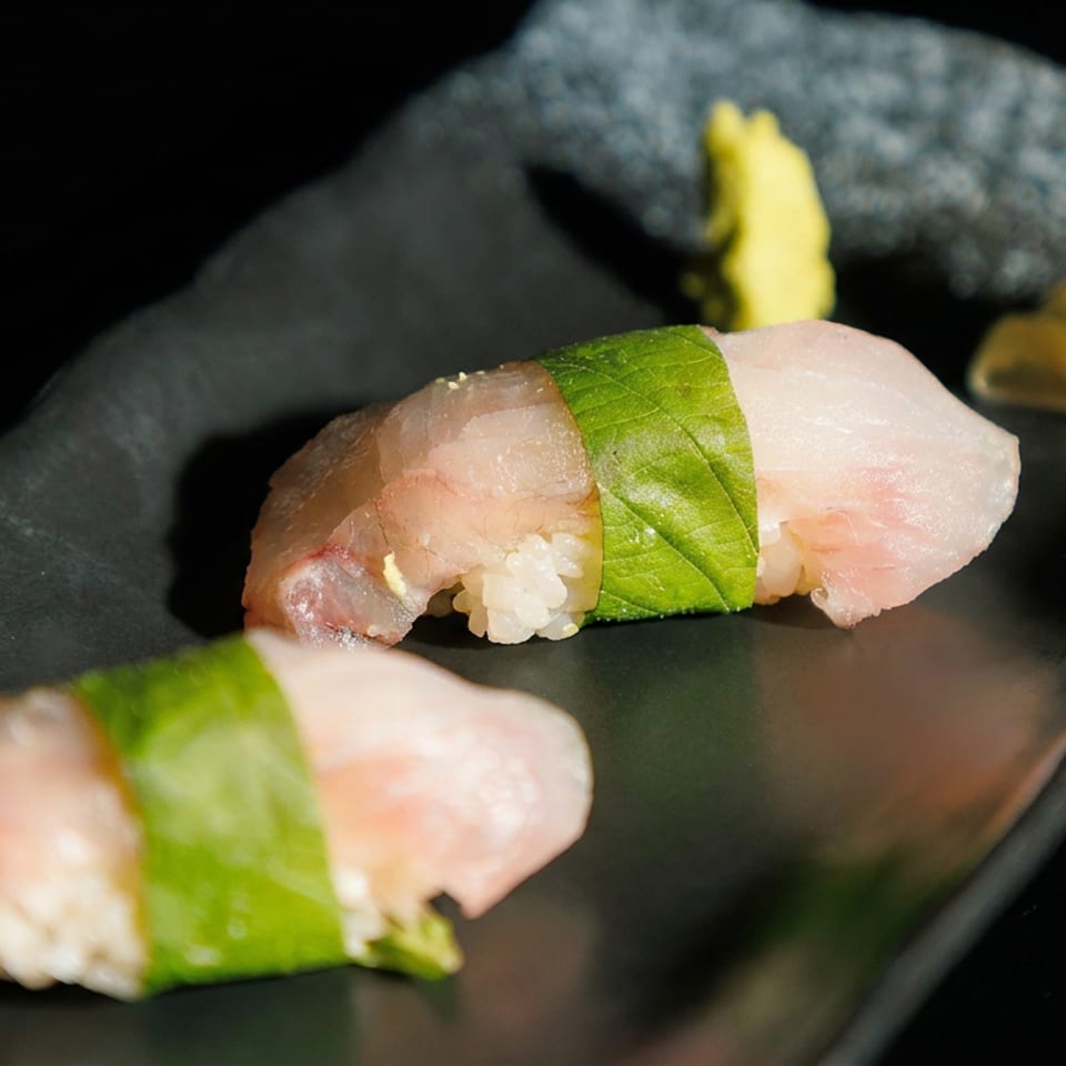 Two pieces of fish sushi wrapped with green leaves sit on a black plate alongside pickled ginger and garlic.
