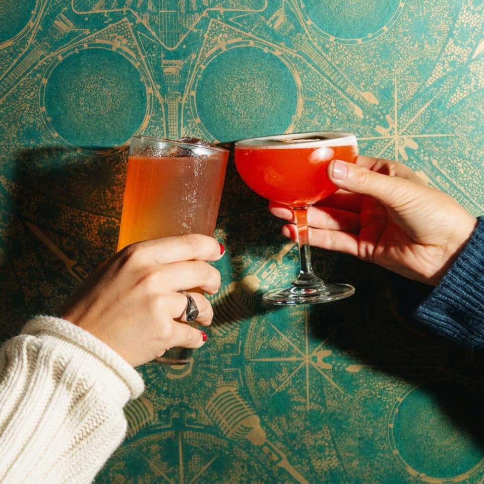 Two people toast with orange and red cocktails against a patterned teal wall in warm sunlight.